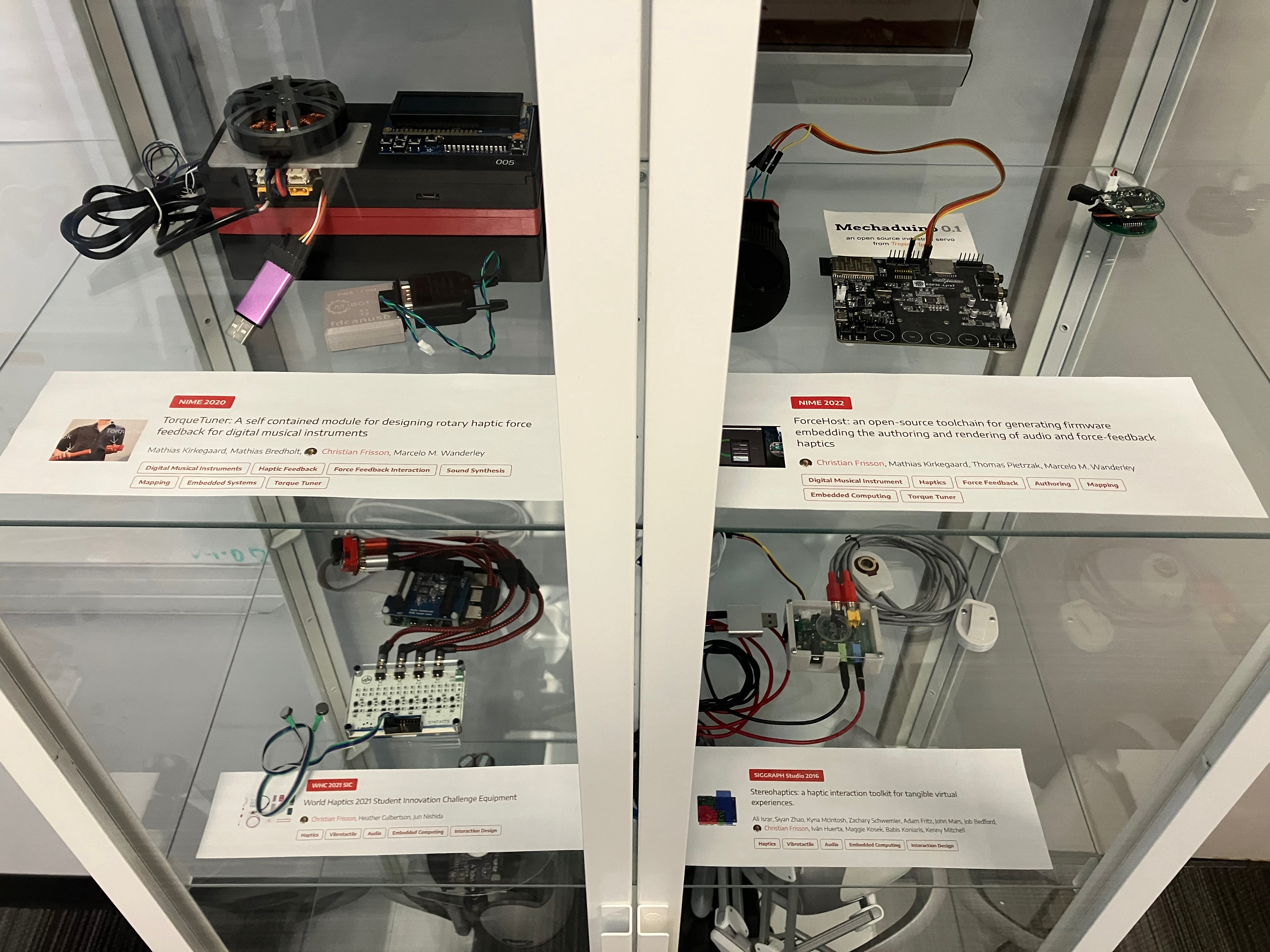 A display case with haptic musical artifacts: force-feedback on top shelf (TorqueTuner and ForceHost), vibrotactile on lower shelf (Syntacts and StereoHaptics).